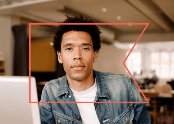 A man looks confidently at the camera. His laptop is open on his desk.